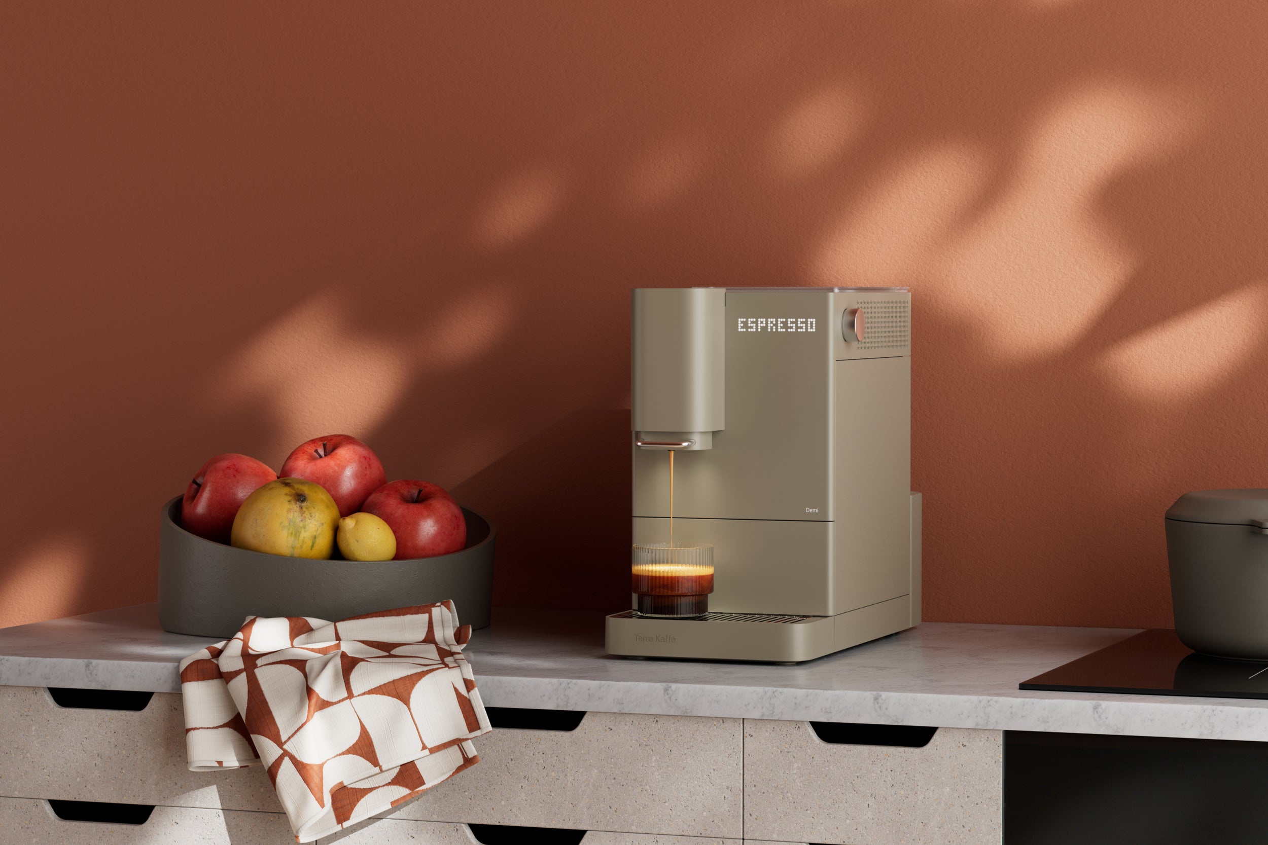 Demi Coffee machine on a kitchen counter with a bowl of fruit and a pot in the background.