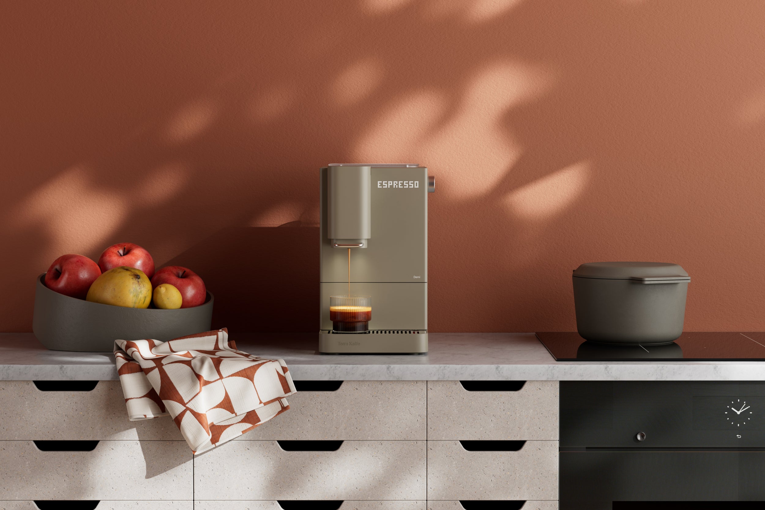 Demi Coffee machine on a kitchen counter with a bowl of fruit and a pot in the background.